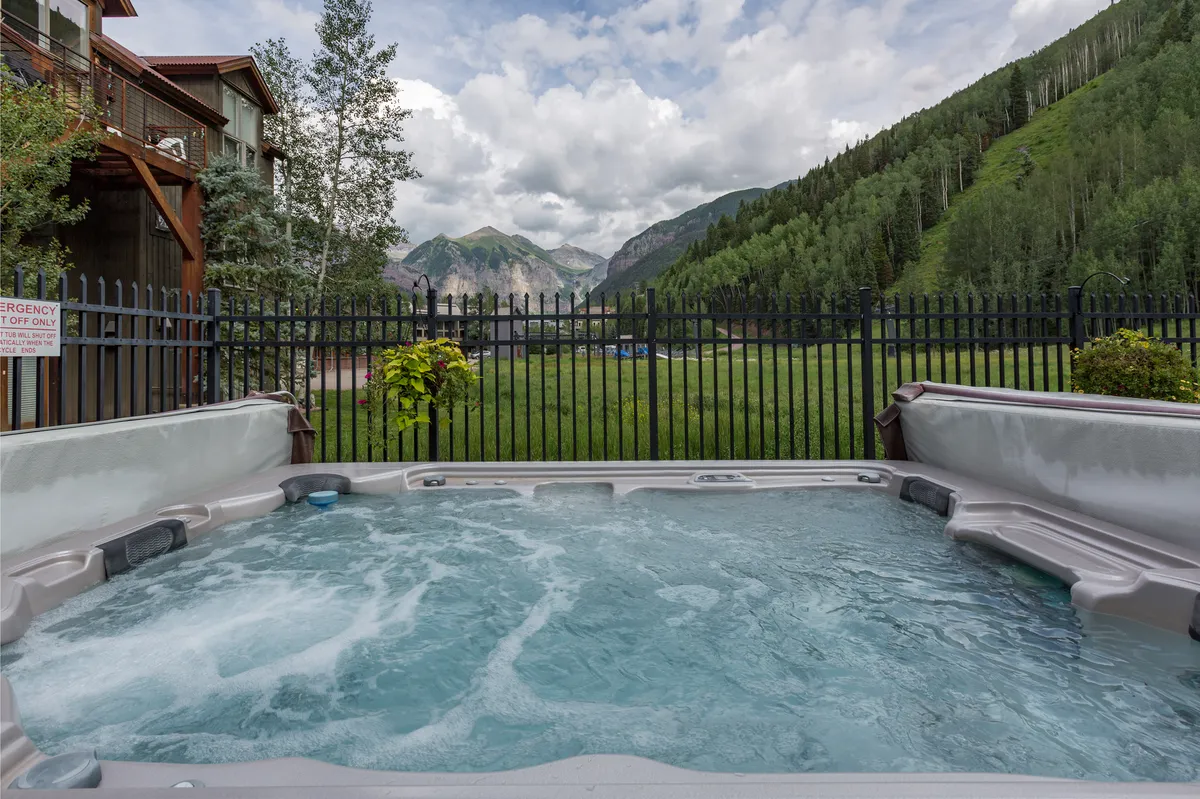 The image showcases an outdoor heated pool with a backdrop of mountains, surrounded by greenery and a fenced area. The mood is serene and rejuvenating, perfect for relaxation after a day of outdoor activity. The pool area is private yet immersed in nature, with opportunities for socializing or simply soaking in the scenery.