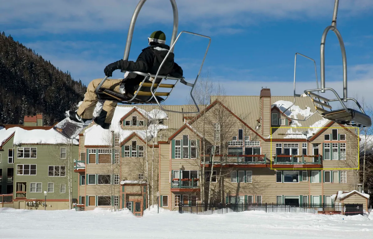 A vibrant winter scene with a ski lift and snow-covered buildings framed by trees, all set against a backdrop of bright blue skies. The image conveys lively activity at the base of the slopes and the inviting atmosphere of a mountain resort.