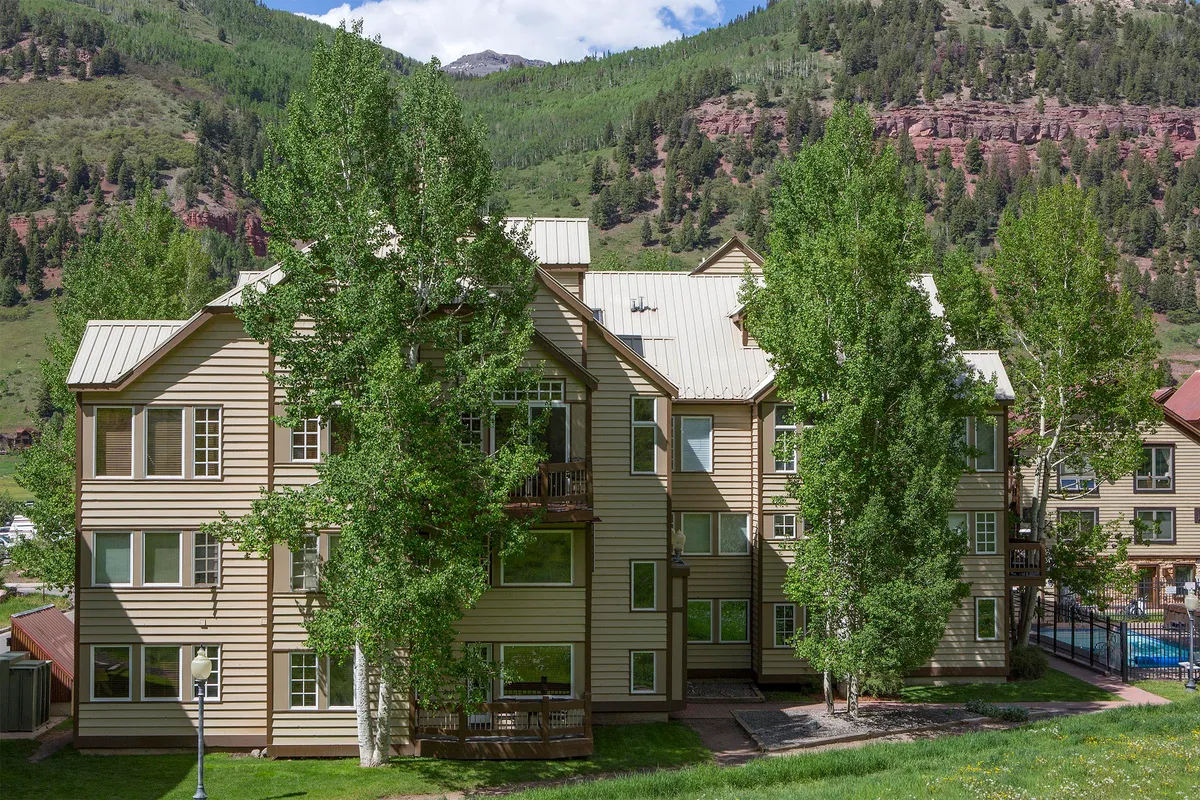 This image features the exterior of a three-story building nestled among lush green trees with a majestic mountain backdrop. The scene exudes a peaceful and serene mood, and hints at activities like hiking, relaxing outdoors, and enjoying sunset views over the mountains. The private balcony is visible, with opportunities for morning coffee or photography against the scenic setting.