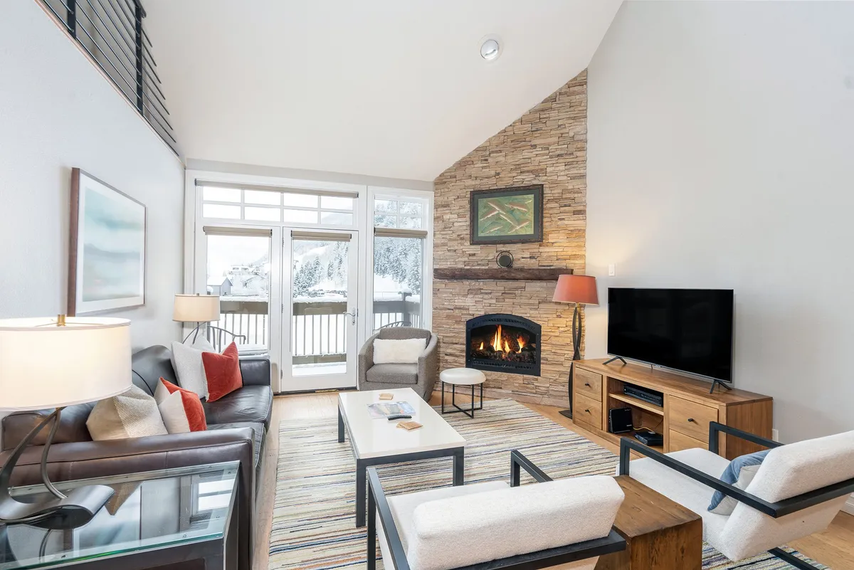 The image showcases a spacious, inviting living area with a roaring fireplace, plush seating arranged for gathering, and expansive windows revealing snowy mountain views. Warm lighting and tasteful mountain-inspired artwork add to the cozy, upscale ambiance.