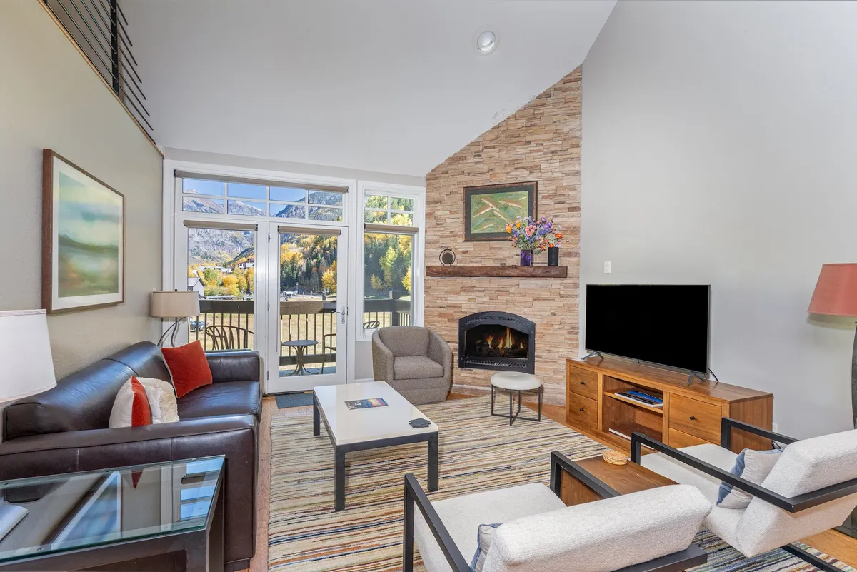 A spacious living room with a stone-faced gas fireplace as the focal point. Large picture windows provide sweeping views of the surrounding mountains and flood the space with natural light. Plush sofas are arranged around the fireplace and flat-screen TV, creating a welcoming gathering space. Artwork and tasteful decor enhance the modern mountain aesthetic.