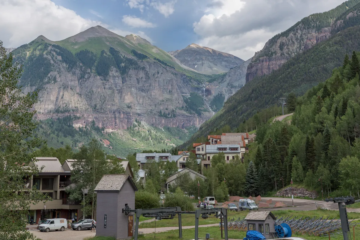 A vibrant Telluride street lined with colorful buildings and lush greenery, framed by majestic mountains under a clear sky. A ski lift hints at year-round adventure, while the architecture and setting evoke the unique charm of the mountain town.