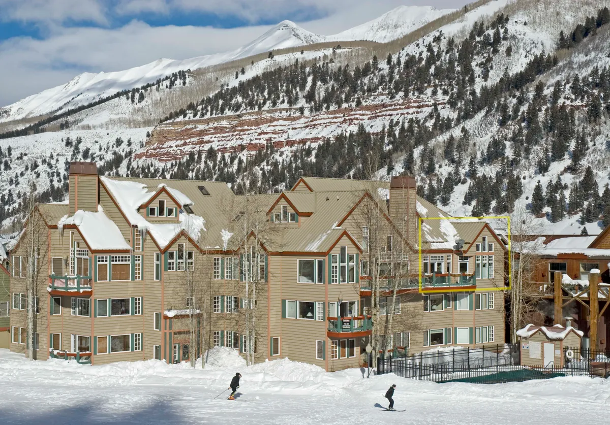 A wide view of the main building in winter, surrounded by snow with people skiing on the nearby slopes. The architecture and balconies are prominent, underscoring ski-in/ski-out access. The lively yet serene mood captures both the thrill of direct ski access and the inviting atmosphere of the complex.