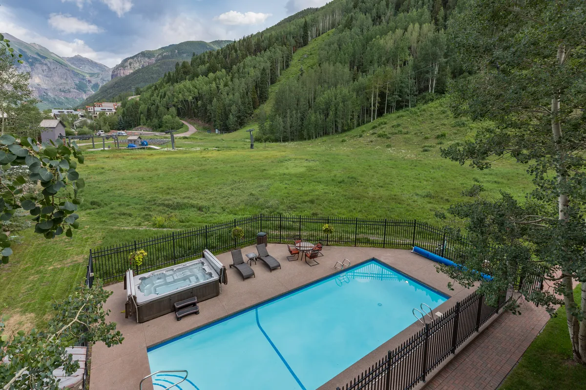 This exterior image showcases the vibrant swimming pool and hot tub area framed by lounge chairs and a picturesque mountain backdrop. Lush greenery surrounds the scene, evoking a tranquil yet active resort atmosphere perfect for relaxation and socializing. The view hints at both summer and year-round enjoyment, with inviting water features and spaces for communal gatherings.