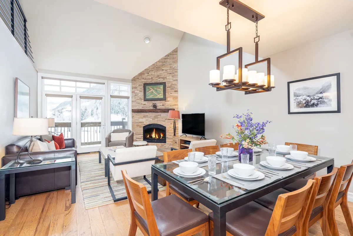 A warm, inviting dining and living area with a set dining table, comfortable seating area, and a prominent fireplace. The space exudes togetherness and hospitality, with balcony access visible, hinting at the transition between indoor gathering and outdoor mountain views.
