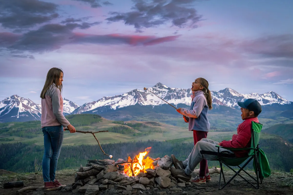 A joyful scene around a campfire with children roasting marshmallows and a parent sitting nearby, all set before a stunning mountain landscape at sunset. The atmosphere is warm and communal, evoking family bonding and memorable outdoor experiences.