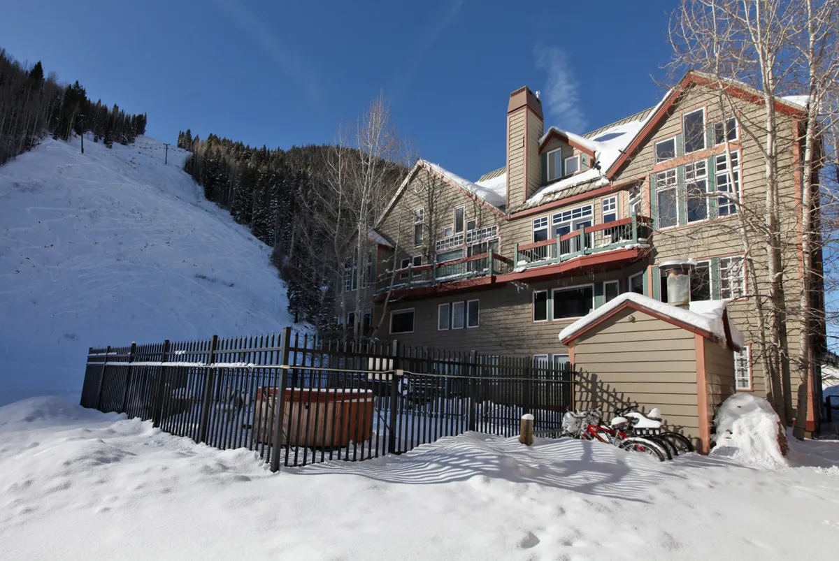 This image captures the building's facade blanketed in snow, with a ski slope directly adjacent and a steaming hot tub in the foreground. Bicycles are also visible, adding a touch of year-round adventure. The snow-covered landscape and proximity of the building to the action highlight direct slope access and après-ski relaxation opportunities.