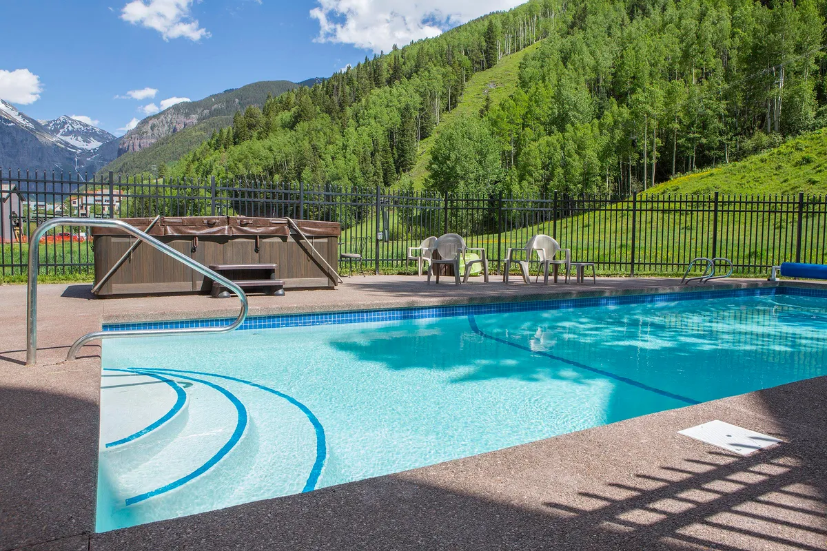 A tranquil exterior shot of the swimming pool and hot tub set against a mountain backdrop and green hillside. Lounge chairs are arranged poolside, inviting relaxation and social gatherings, while the open space provides opportunities for both summer fun and year-round enjoyment.