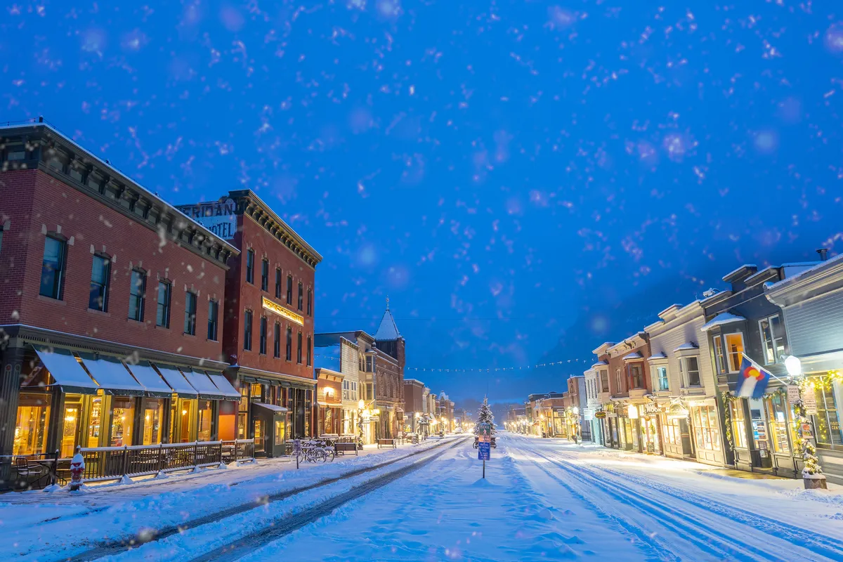 A picture-perfect, snow-covered street adorned with festive decorations, colorful buildings, and a sparkling Christmas tree. Streetlights cast a warm glow, and the scene radiates winter charm, inviting guests to stroll into town for seasonal festivities and cozy cafés.