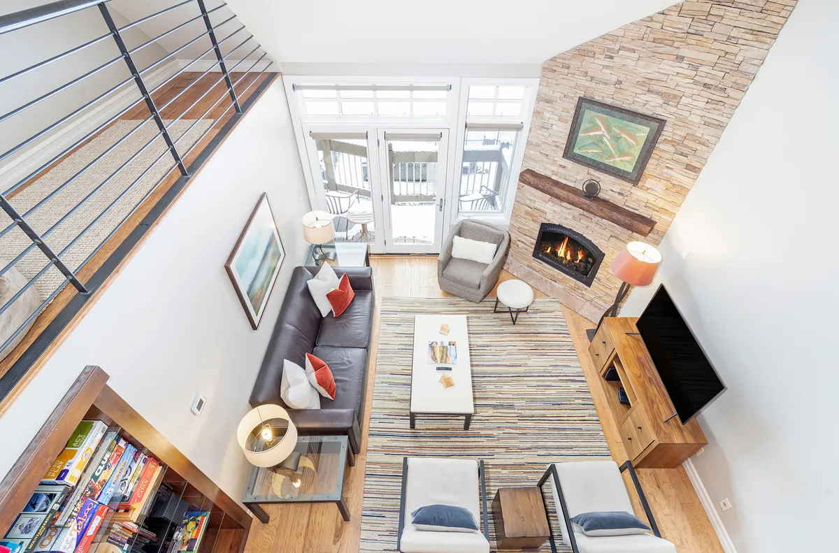A modern, cozy living area featuring a stylish sofa, coffee table, large windows with abundant natural light, and a stone-faced fireplace. The open layout seamlessly connects the living area to the rest of the home, with views of the outdoors visible through the expansive windows.